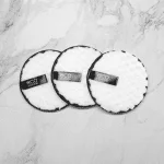 Three round, white Reusable Makeup Remover Pads - Black with black trim are laid out on a marble surface. These pads, labeled "eco essentials," offer an eco-friendly alternative for your skincare routine.